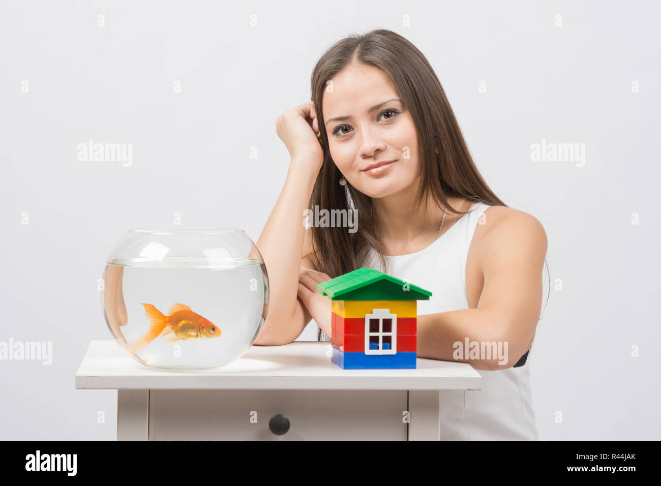 La ragazza si siede a un tavolo su cui vi è un acquario con pesci rossi e toy house Foto Stock