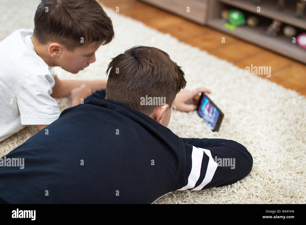 Ragazzi piccoli guardando un video tramite smartphone su un tappeto a casa, vista posteriore Foto Stock