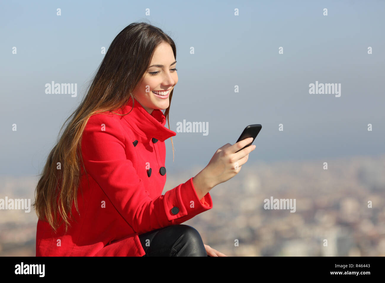 Felice ragazza in rosso con uno smart phone in inverno con la città in background Foto Stock