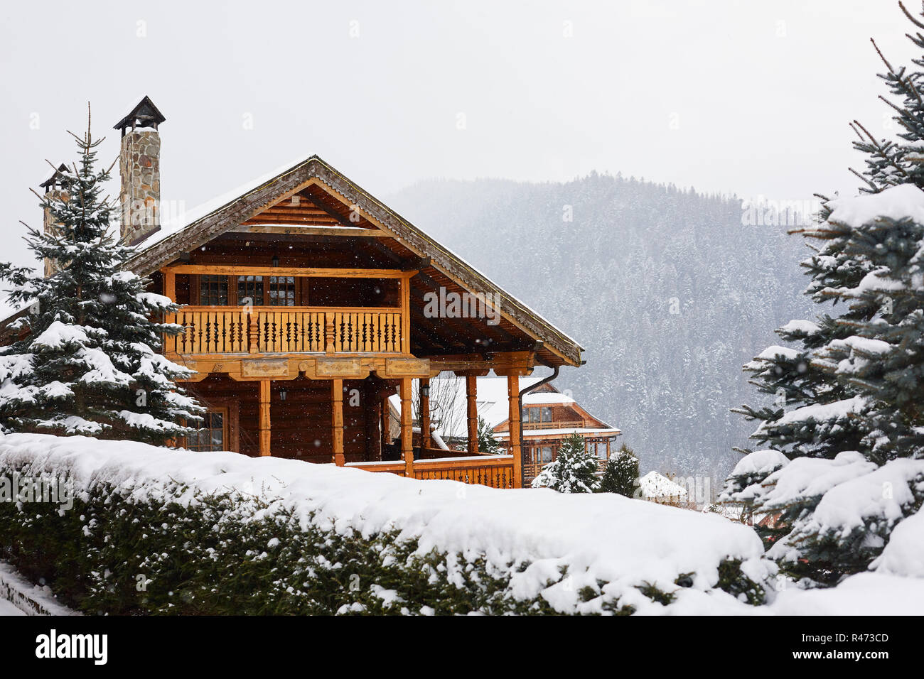 Natale mansion di legno in montagna su nevicata giorno d'inverno. Accoglienti chalet sulla stazione sciistica vicino alla foresta di pini. Cottage di legno tondo con balcone in legno. Abeti coperti di neve. Camini di pietra. Foto Stock