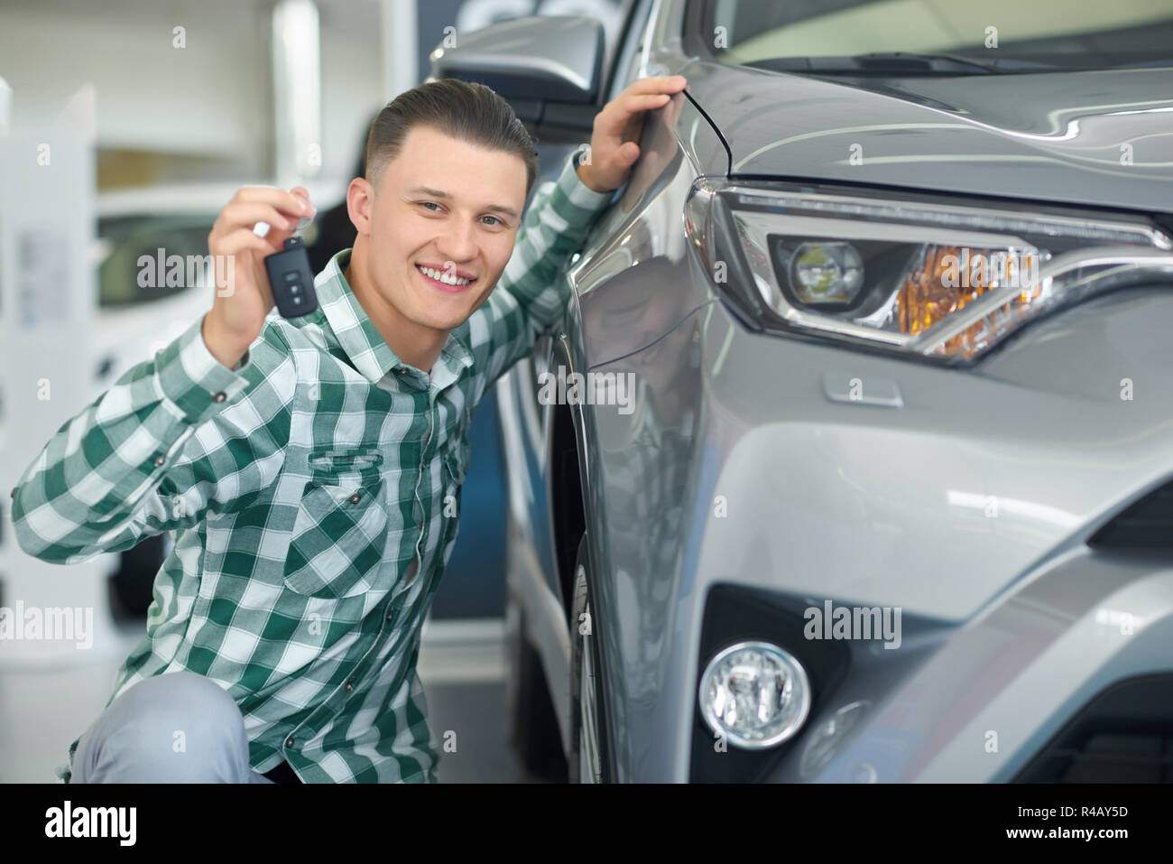 Attraente client bionda la scelta di auto e rannicchiati vicino a uno di lusso. Chiusura del bel giovane proprietario di grigio grandi auto tenendo la sua mano su di esso. Sorridenti conducente veicolo di acquisto e che mostra la chiave. Foto Stock