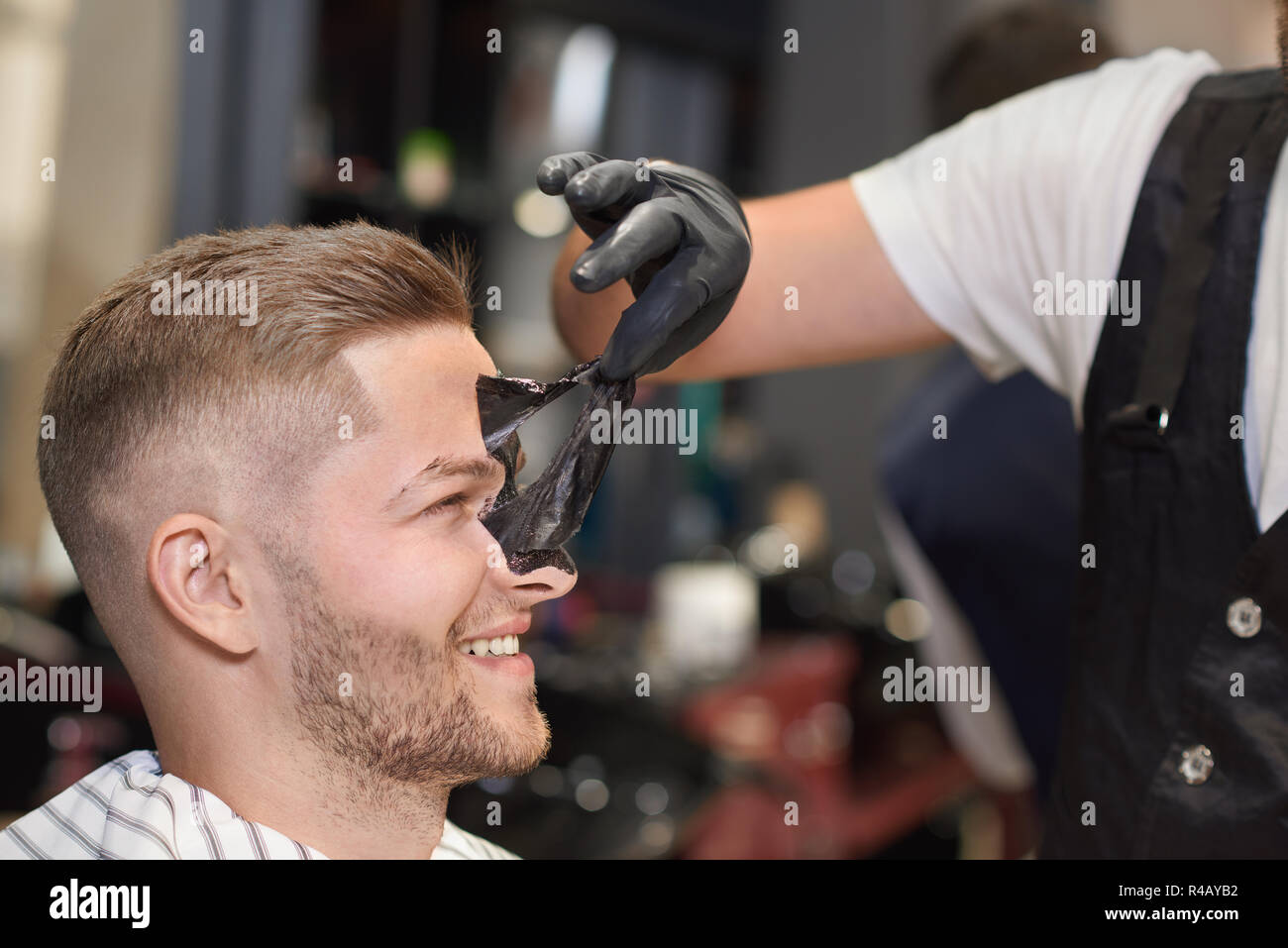 Vista laterale del processo di rimozione di maschera nera dalla faccia del client maschio in un barbiere. Barbiere in guanti di protezione tenendo lontano maschera contro i punti mentre il cliente a ridere. Concetto di cura. Foto Stock