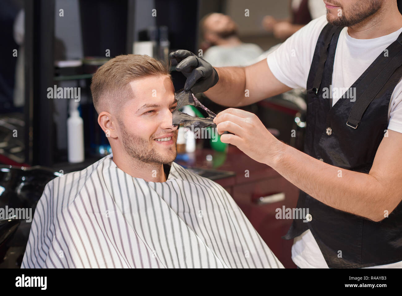 Uomo sorridente seduto su una sedia in un barbiere mentre il barbiere in guanti protettivi rimozione maschera nera dalla faccia del client. Uomo bello facendo pulizia procedura contro i puntini. Concetto di cura. Foto Stock