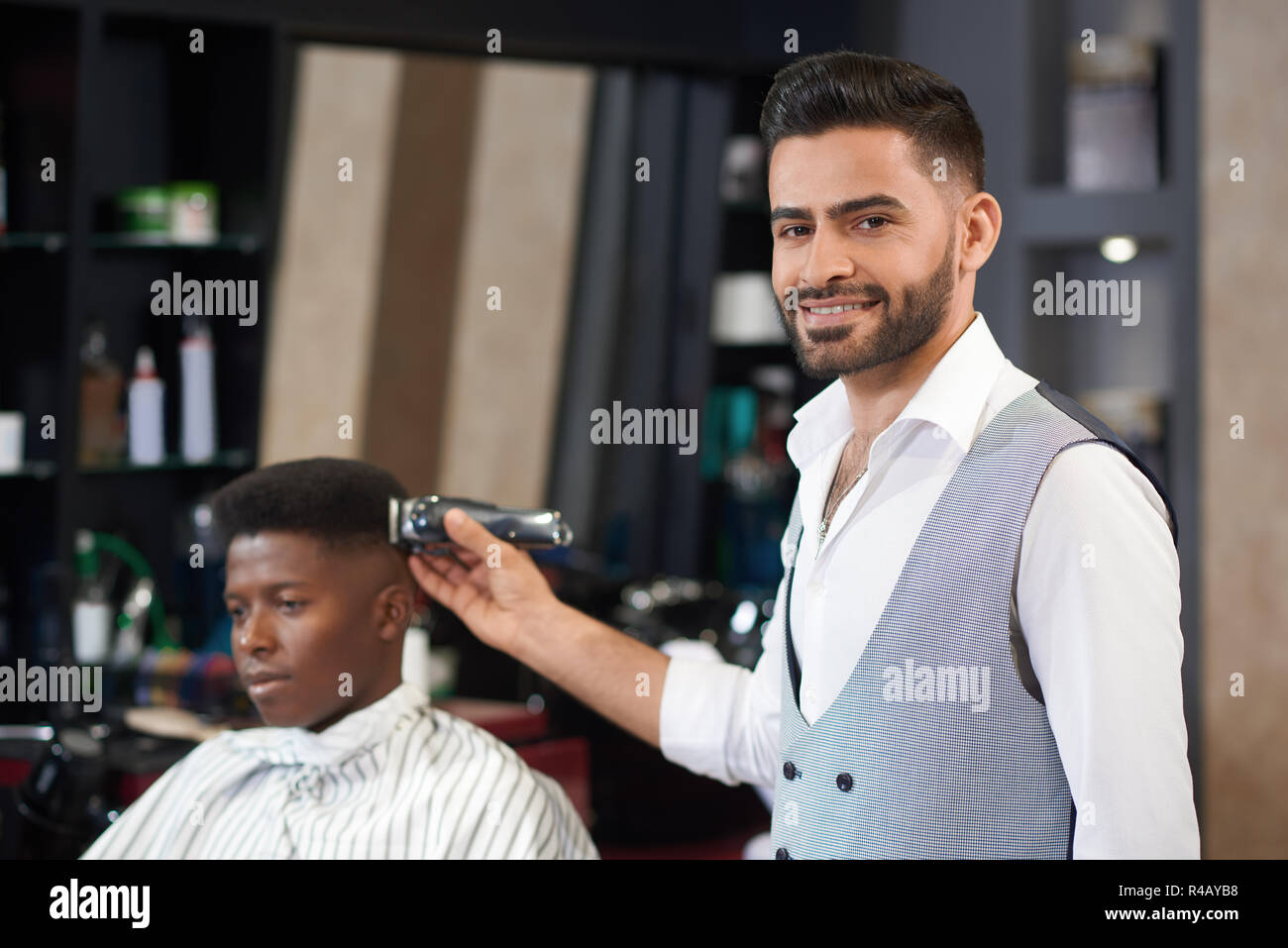 Vista frontale del barbiere elegante in una camicia bianca e gilet guardando la telecamera, in posa e sorridente in un barbiere. Parrucchiere maschio facendo i capelli di fresatura con clipper al cliente seduto a sfondo. Foto Stock