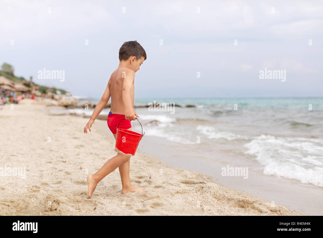 Ragazzo sulla spiaggia a piedi al mare portante una benna, Grecia Foto Stock