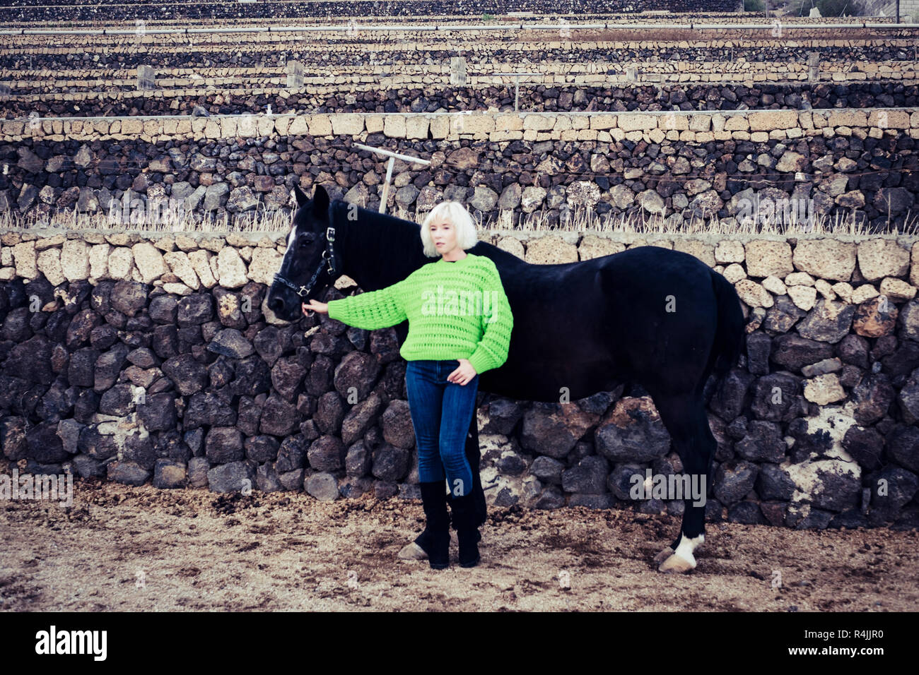 Il concetto di moda con grazioso modello bionda in posa in campagna con un cavallo nero e indossa una giacca verde. i colori e il contrasto delle persone e degli animali Foto Stock