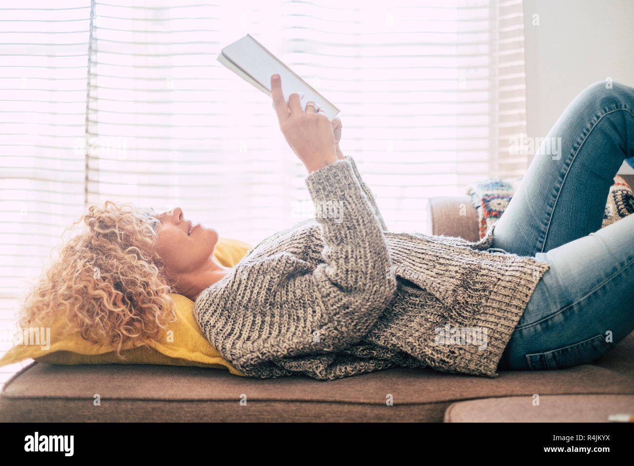 Bella la mezza età femmina di deporre sul divano di casa a leggere un libro e godendo la luce dall'esterno. Persone appoggiano e prendere una pausa dopo il lavoro - Foto Stock