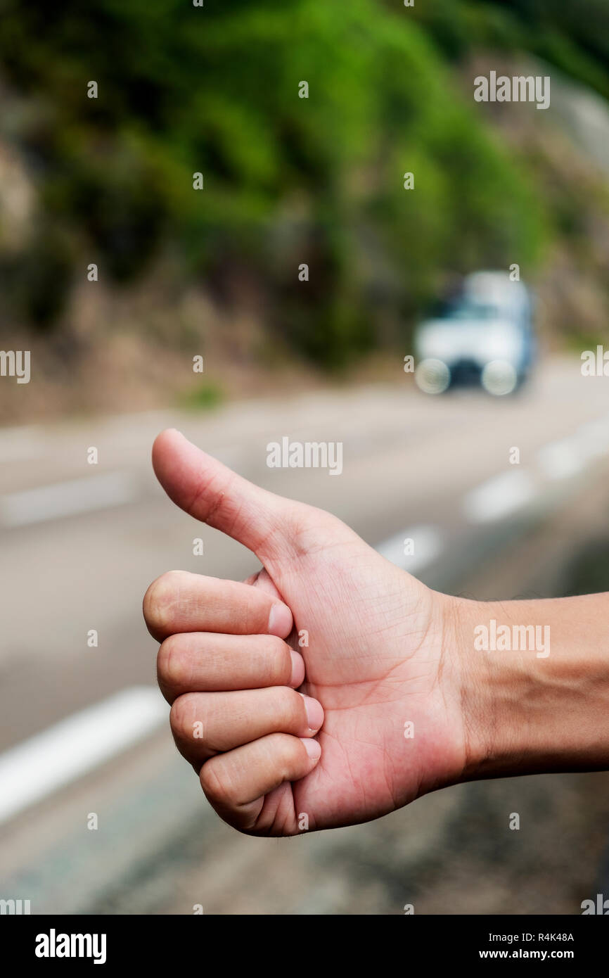 Primo piano di un giovane uomo caucasico autostop in piedi sul ciglio della strada di una strada secondaria con il suo pollice in alto Foto Stock