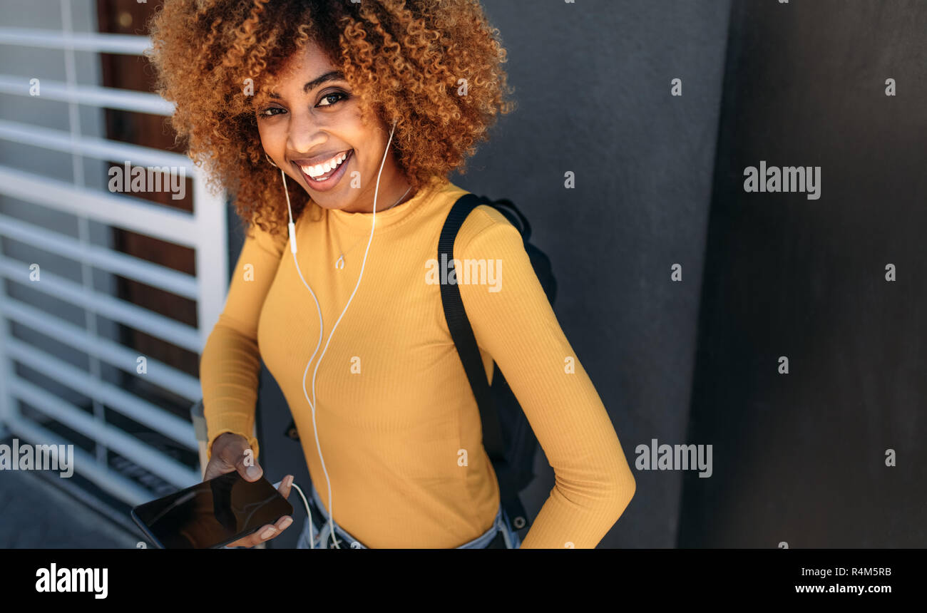 Sorridente afro american turista femminile indossando uno zaino ascoltando la musica sul telefono cellulare. Donna allegra ascoltando musica su auricolari tenendo un MOB Foto Stock