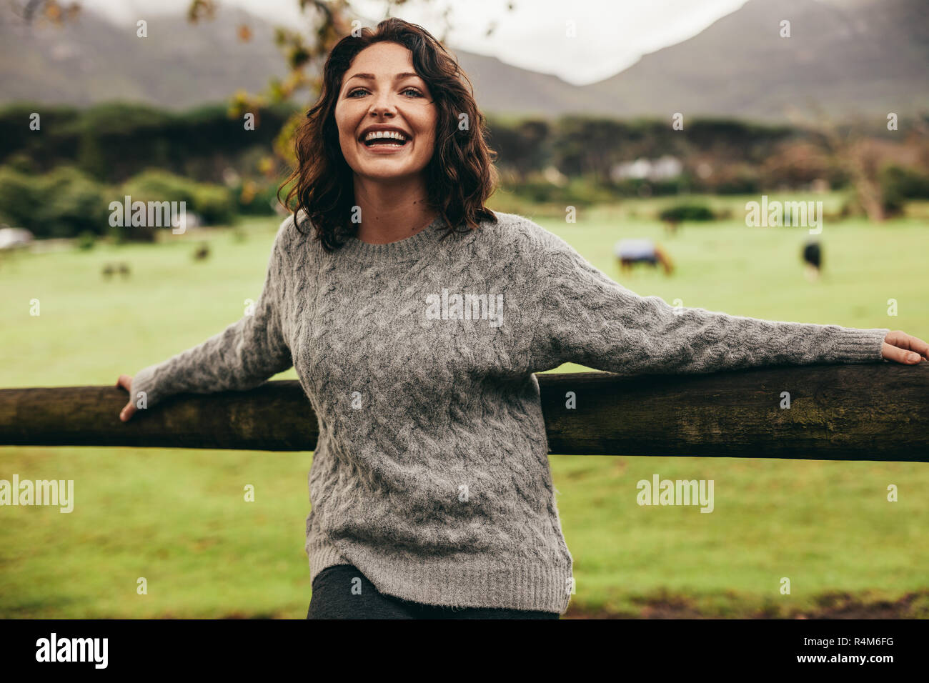 Ritratto di donna allegra appoggiandosi a un recinto in campagna. La donna che indossa un maglione in piedi da una recinzione guardando la fotocamera e ridere. Foto Stock
