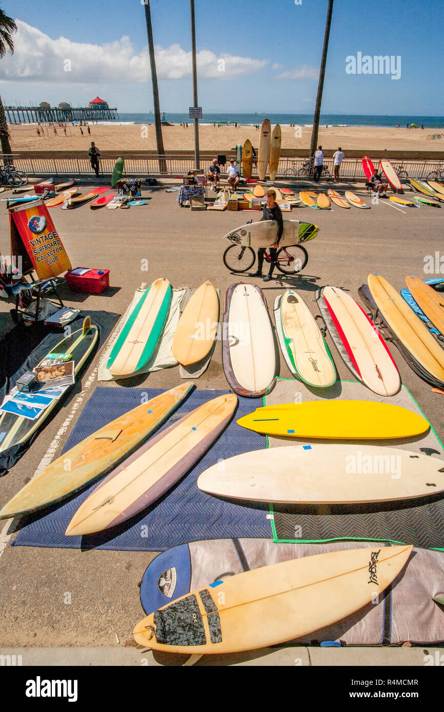 Toting una piccola tavola da surf, un ciclista si affaccia su di una mostra di vintage tavole lunghe sotto Pier Plaza a Huntington Beach, CA. Nota pier in background. Foto Stock