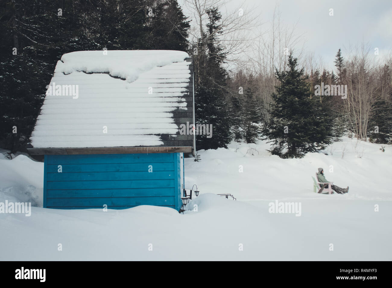 Una donna che si rilassa in una sedia adirondack di fronte alla sua piccola casa blu nella neve Foto Stock