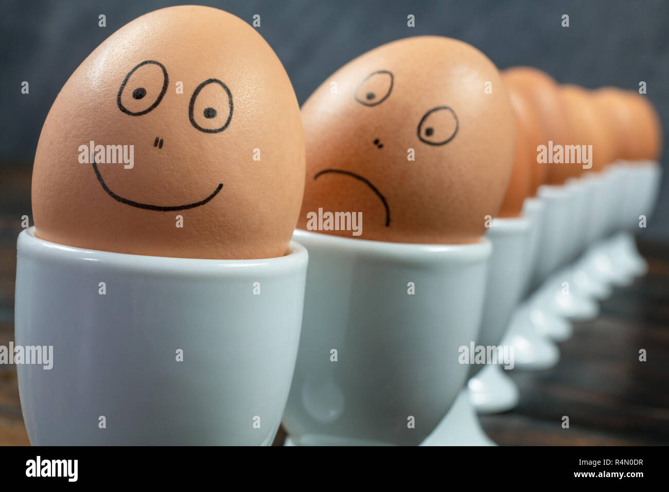 Concetto di coda di attesa o la linea fino a dieci uova sode in bianco d'uovo con coppe disegnate felici e tristi si affaccia su di un tavolo di legno Foto Stock