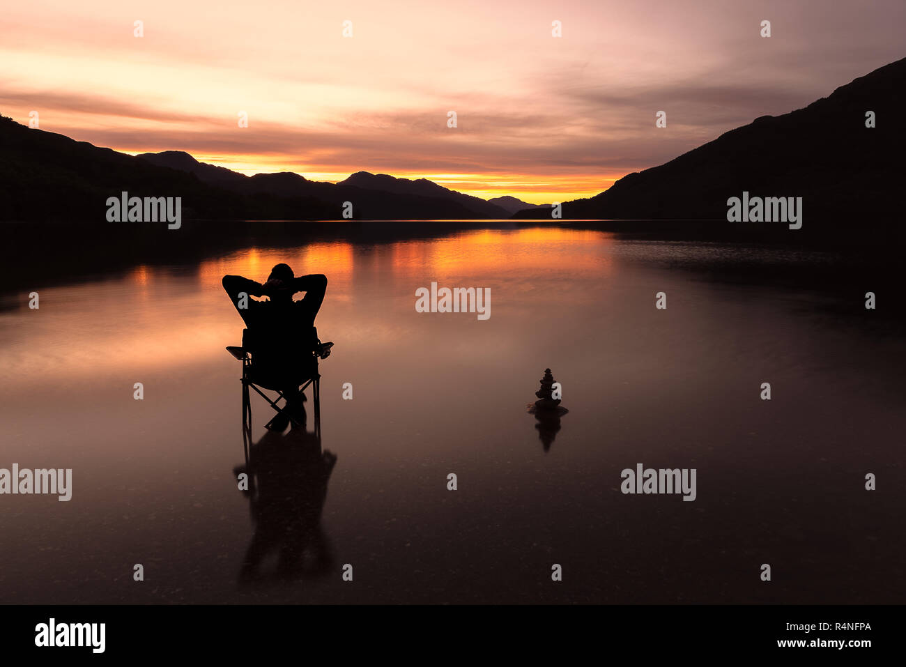 Lone figura, uomo, seduta su una poltrona rilassante, accanto alla pila di pietra, come i tramonti sul Loch Lomond Scozia, Regno Unito Foto Stock