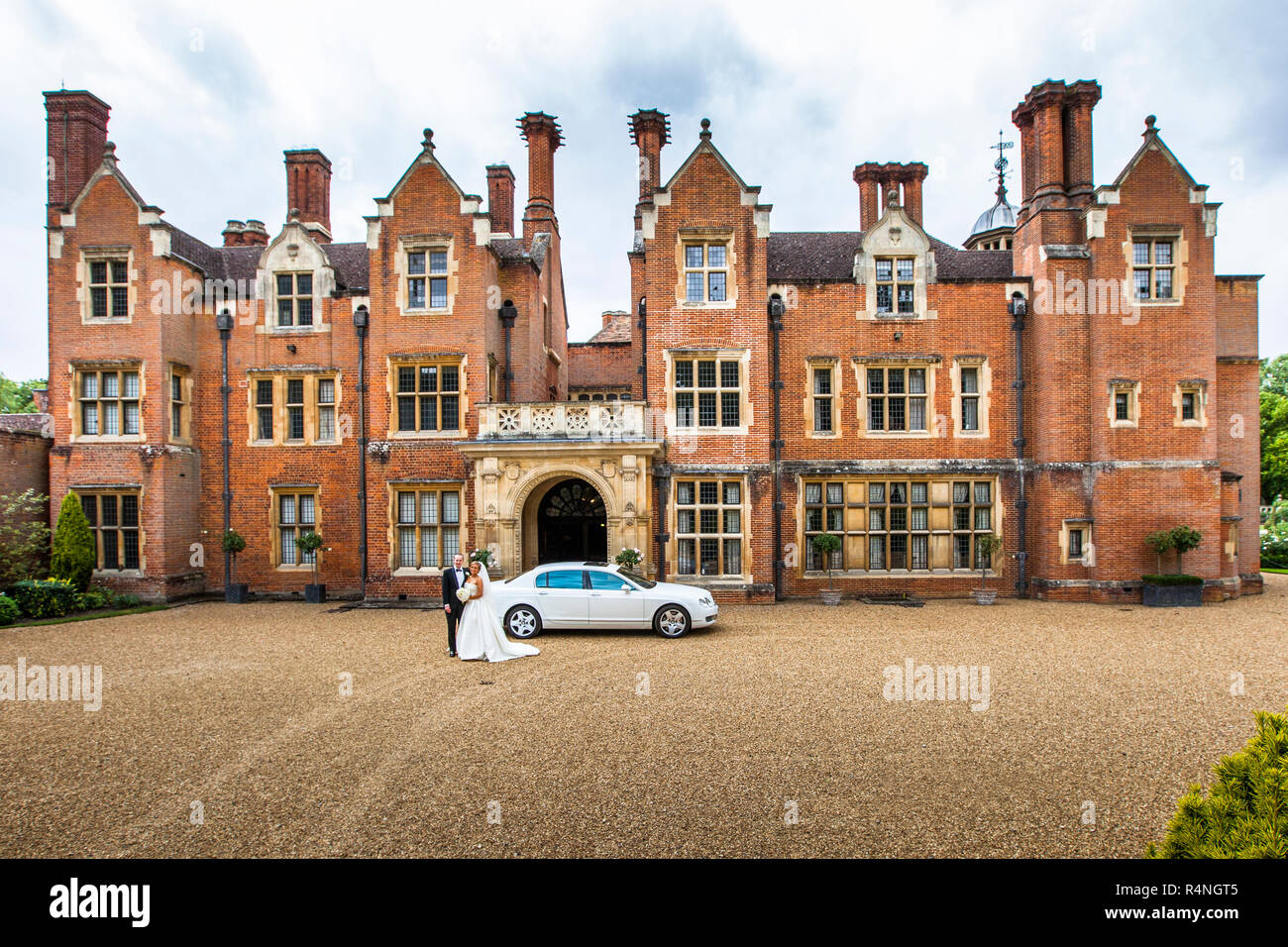 La coppia di sposi posa davanti alla facciata in mattoni rossi di Longstowe Hall vicino a Cambridge, in Inghilterra, accanto a un'auto da matrimonio bianca. La residenza fu costruita alla fine del XVI secolo in stile elisabettiano e fu ristrutturata tra il 1907 e il 1914 dall'architetto Sir John Simpson in stile giacobino. Foto Stock