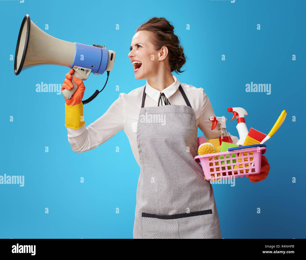 Felice cameriera nel grembiule con cestello con detergenti e spazzole urlando attraverso il megafono isolato sul blu. Attenzione! - Semplice e senza problemi di CLE Foto Stock