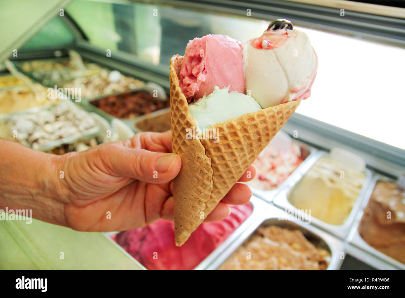 Mano femminile è in possesso di una grande fragola, menta, vaniglia con ciliege acide di gelato in cono di cialda. Gelato frigo con servizio di acciaio contenitori. Foto Stock