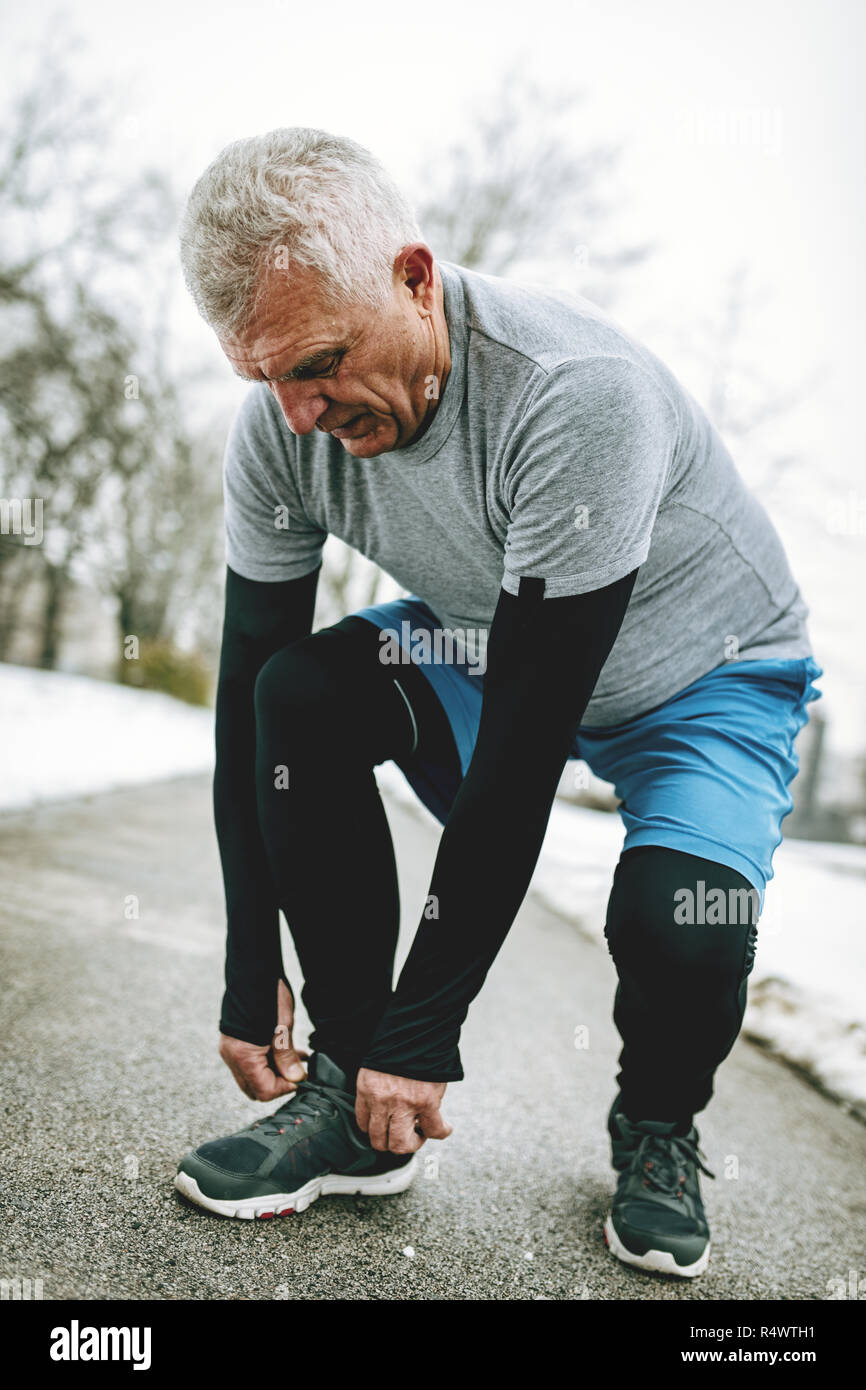 Active senior legatura di scarpe da corsa e fare una pausa durante il jogging e fare gli esercizi nel parco pubblico in inverno di formazione al di fuori. Foto Stock