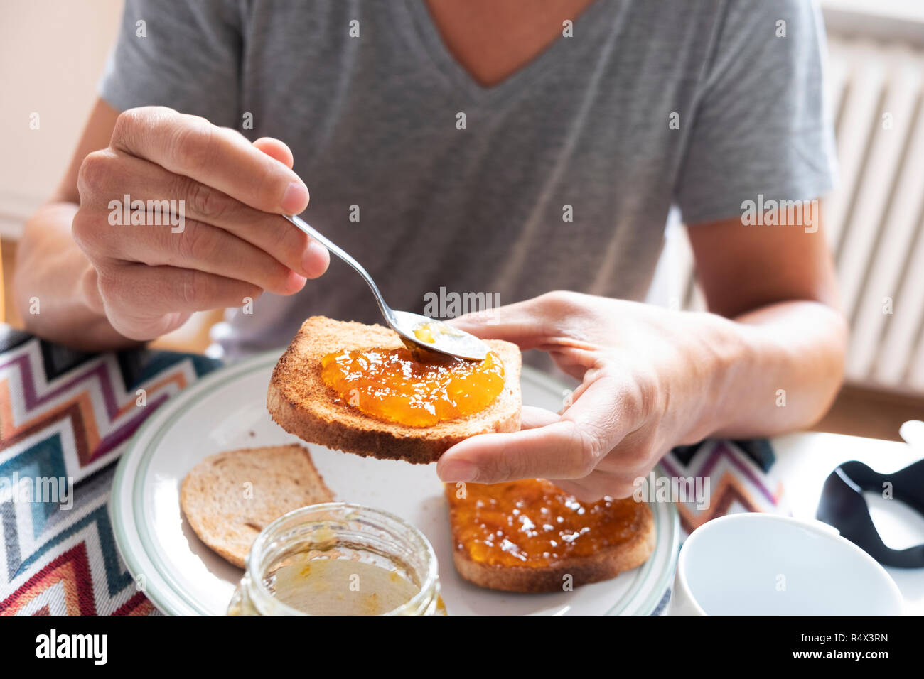 Primo piano di un giovane uomo caucasico, indossando un grigio casual T-shirt, seduti a un tavolo set, diffondendo alcuni arancione o confettura di pesche su un toast Foto Stock