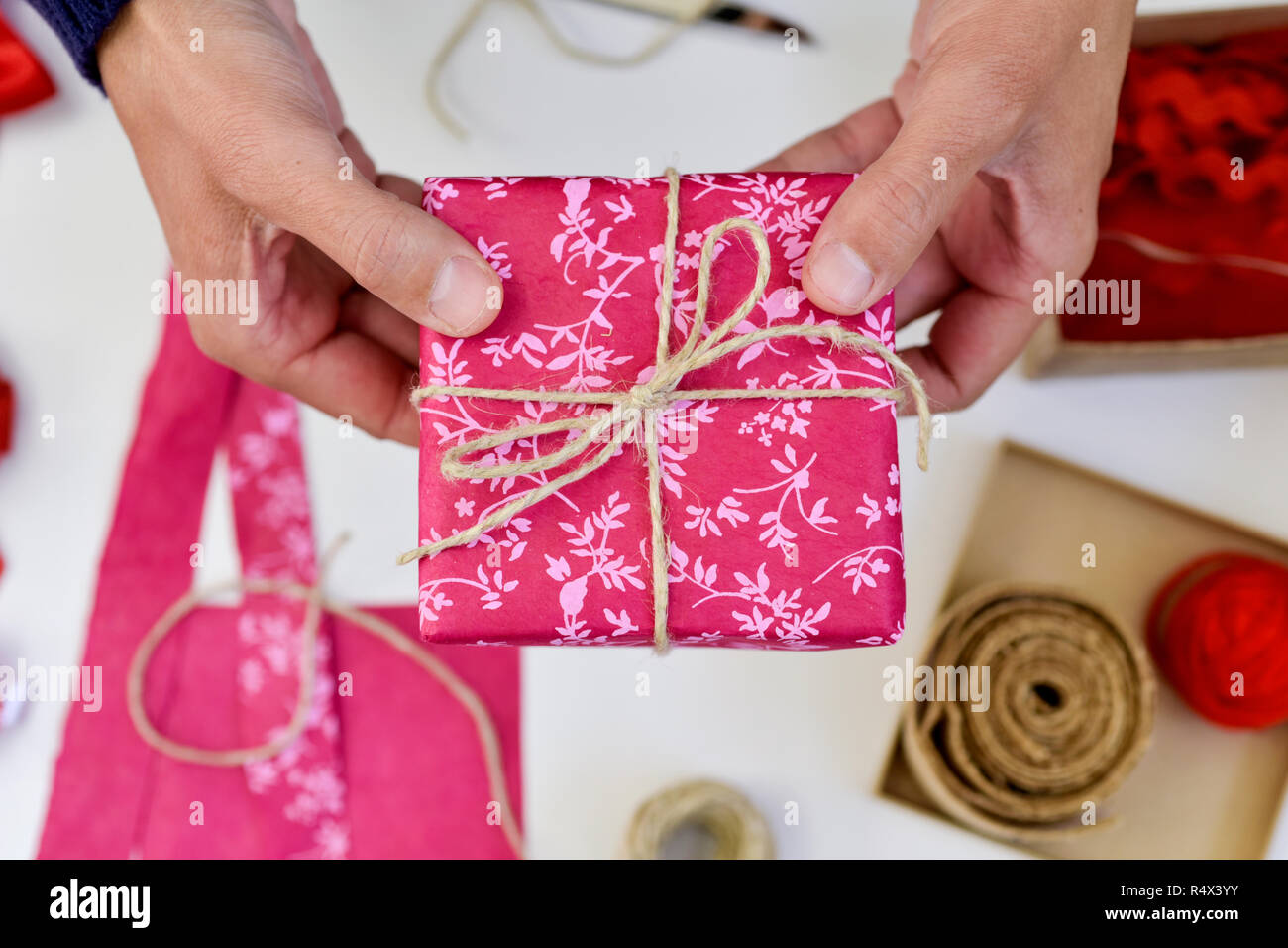 Elevato angolo di visione di un giovane uomo caucasico con un bel regalo in mano, avvolto in carta rosa e legato con un rustico di stringa, su un tavolo bianco pieno di Foto Stock