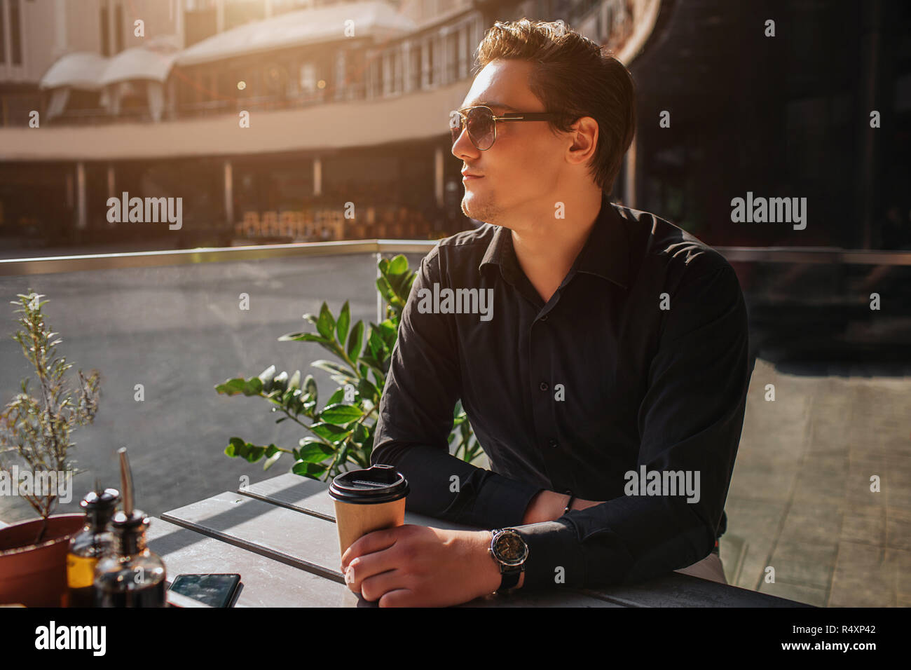 Attraente giovane uomo seduto a tavola e guardare a sinistra. Egli tenere tazza di bevanda. Egli siede all'esterno. C'è il sole. Foto Stock