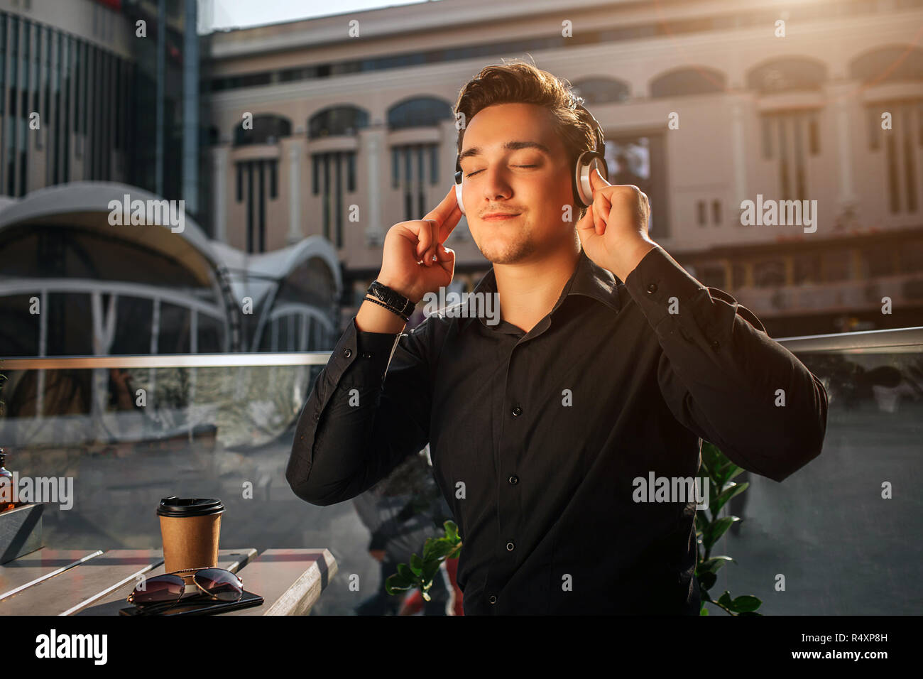 Felice giovane uomo seduto a talbe esterno e ascoltare la musica attraverso le cuffie. Egli enjoyes. Guy tenere le mani sulle cuffie. C'è il sole. Foto Stock