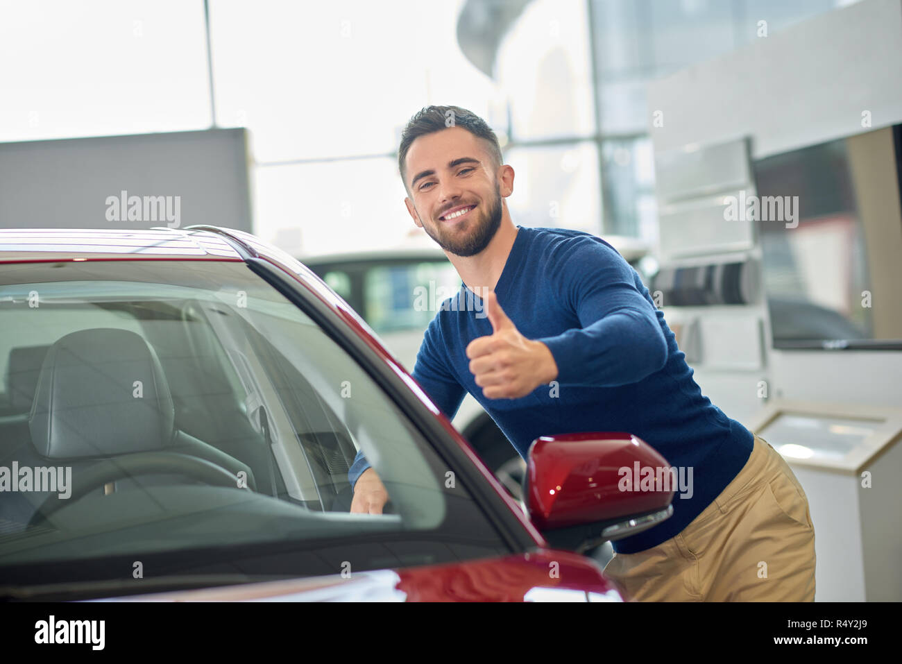 Brunette uomo bello con la barba arrivando in auto salon. Attraente un cliente contento e sorridente in posa vicino al red auto. Il giovane driver in blu maglione appoggiato su auto della porta anteriore e mostra come la. Foto Stock