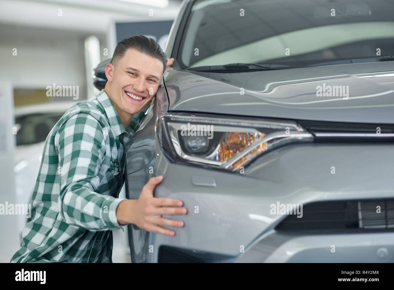 Felice bionda uomo sorridente dopo aver acquistato la sua prima auto. Attraente client di giovani la scelta di auto e accovacciata vicino ad essa. Bello il proprietario in camicia a scacchi davvero gradimento del suo veicolo e avvolgente. Foto Stock