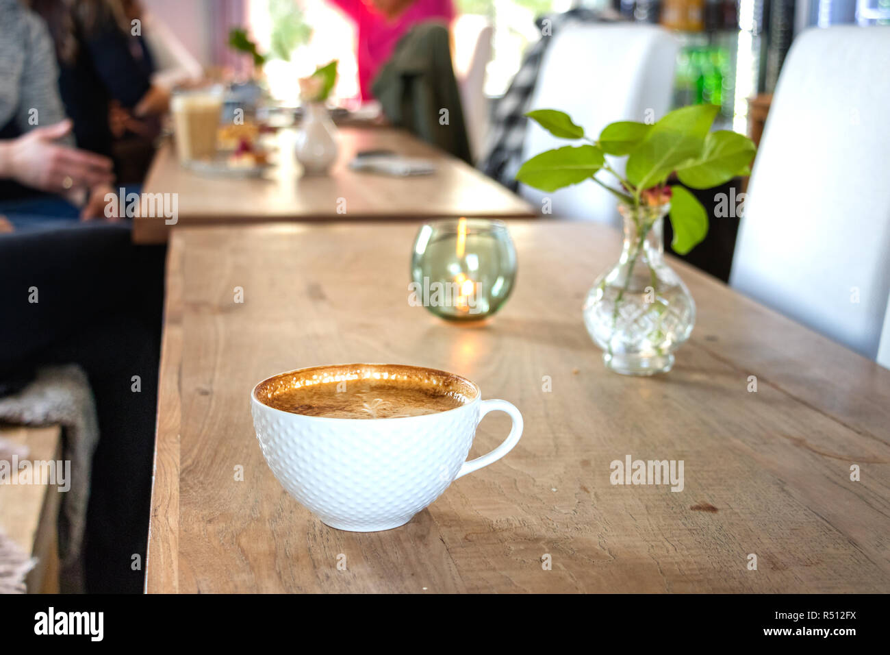 Un bianco tazza di cappuccino su un grande tavolo in legno con persone in background in un coffee shop in Leknes, Norvegia. Foto Stock