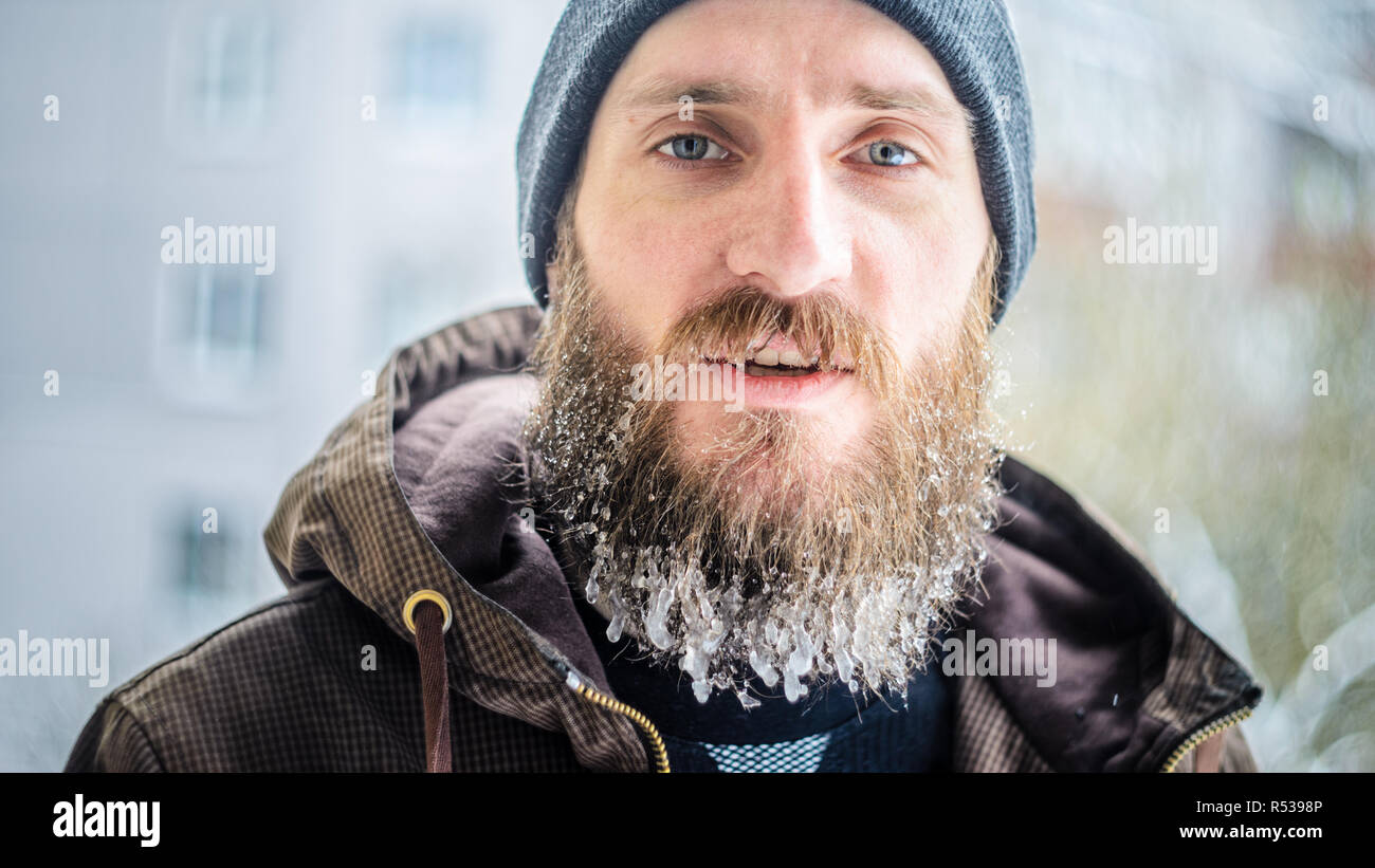 Ritratto di un giovane uomo in grigio di un cappello e una giacca marrone con una lunga barba ricoperta di ghiaccioli. Atleta dopo il jogging all'aperto d'inverno. Inverno Foto Stock