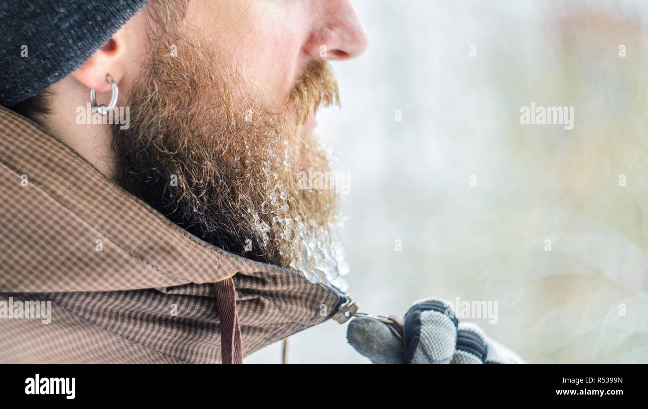 Giovane uomo inverno ritratto nel profilo. La mano guantata zips una camicia sotto il mento la barba ricoperta di ghiaccioli e gocce congelate. Concetto di brina Foto Stock