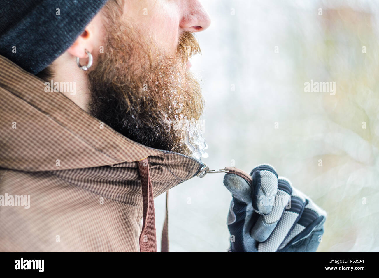 Giovane uomo inverno ritratto nel profilo. La mano guantata zips una camicia sotto il mento la barba ricoperta di ghiaccioli e gocce congelate. Concetto di brina Foto Stock