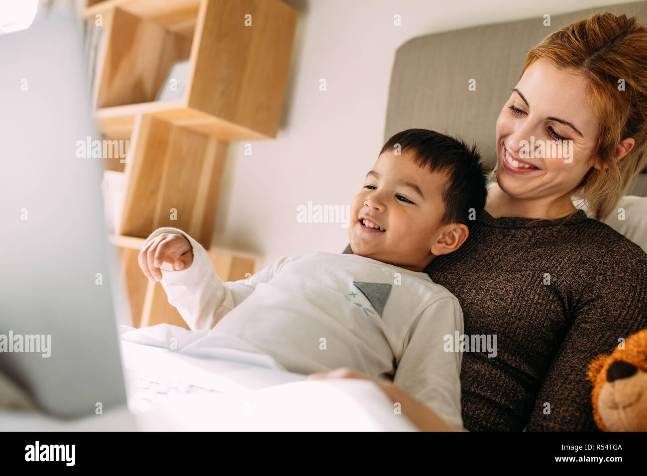 Sorridente giovane donna con un bambino piccolo sul letto per guardare film sul computer portatile. Carino il ragazzo con la madre guardando il laptop. Foto Stock