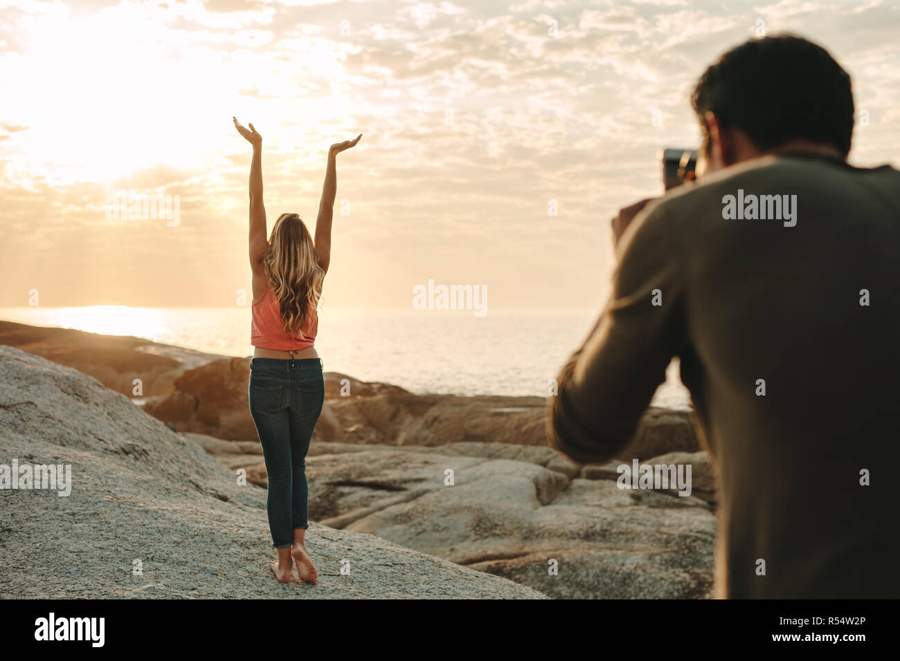 Uomo di prendere una fotografia di una donna in piedi su una roccia presso la spiaggia utilizzando un telefono cellulare. Vista posteriore della donna con le braccia sollevate e in cerca di sole e Foto Stock
