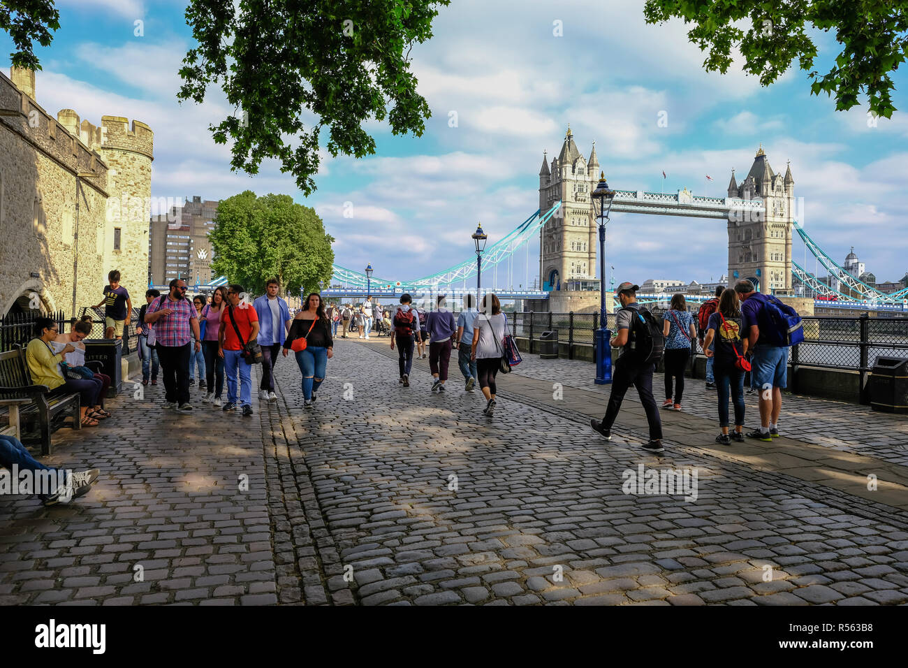 Tower, London, Regno Unito - 8 Giugno 2018: Cobbed pietre e persone che passeggiano accanto alla Torre di Londra con il Tower Bridge in background. Prese su un soleggiato Foto Stock