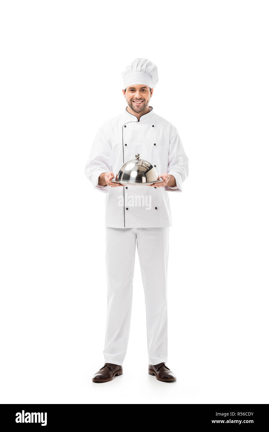 Bel giovane chef holding vassoio con cupola e guardando la telecamera isolato su bianco Foto Stock