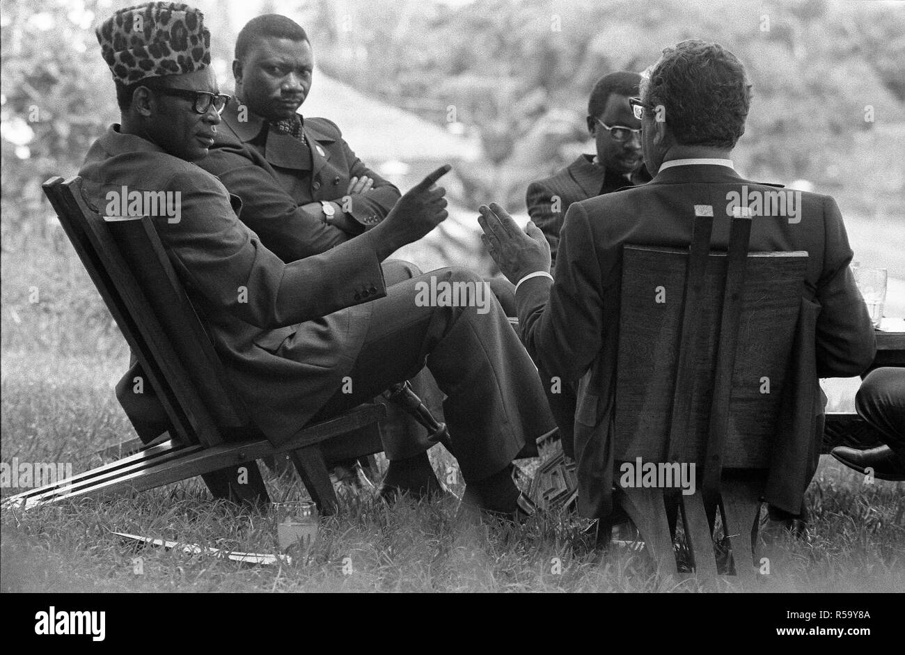 04/28/1976 - la residenza presidenziale - Kinshasa, Zaire - il Segretario di Stato Henry Kissinger A., Presidente Mobutu Sesi Seko, altri - seduti in cerchio su un prato sedie, parlando; Henry Kissinger torna alla fotocamera; Mobutu che indossa a pelle di leopardo hat - Kissinger la diplomazia Viaggio in Zaire e Africa Foto Stock