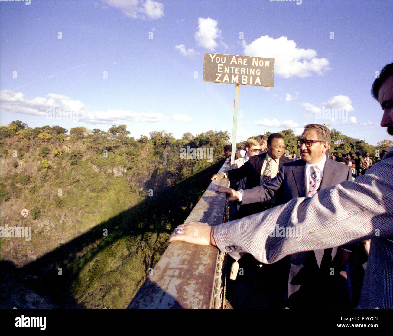 1976, 27 Aprile - Victoria Falls, Zambezi River - Zambia/Rhodesia confine - Henry Kissinger, funzionari dello Zambia, Entourage, Media - in piedi su entrambi i lati di un ponte chiuso su Zambia/Rhodesia frontiera, al di sopra di Victoria Falls; in piedi dal segno - Segretario di Stato Viaggio in Africa; Viaggio a Victoria Falls e Zambia/Rhodesia confine Foto Stock