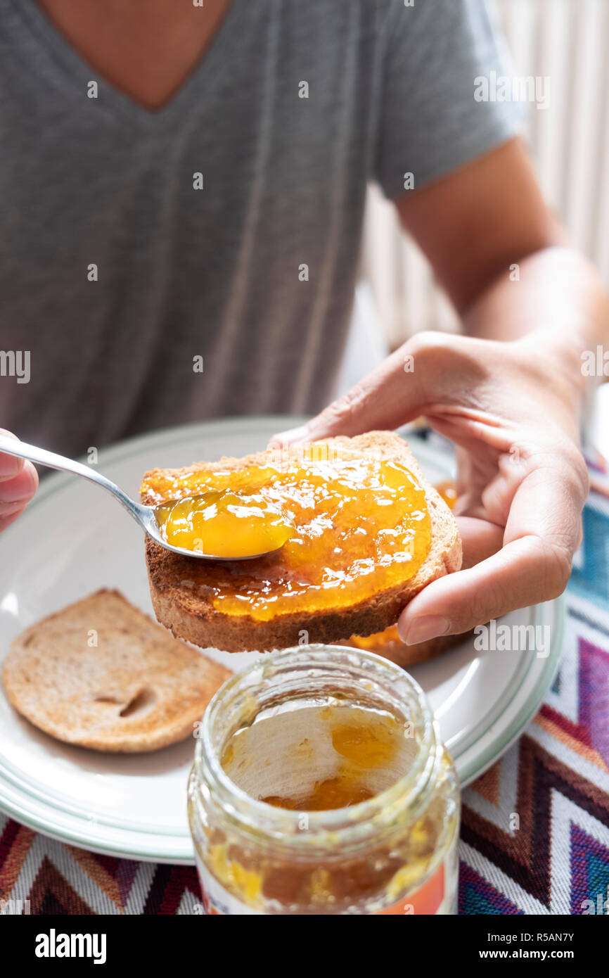 Primo piano di un giovane uomo caucasico, indossando un grigio casual T-shirt, seduti a un tavolo set, diffondendo alcuni arancione o confettura di pesche su un toast Foto Stock