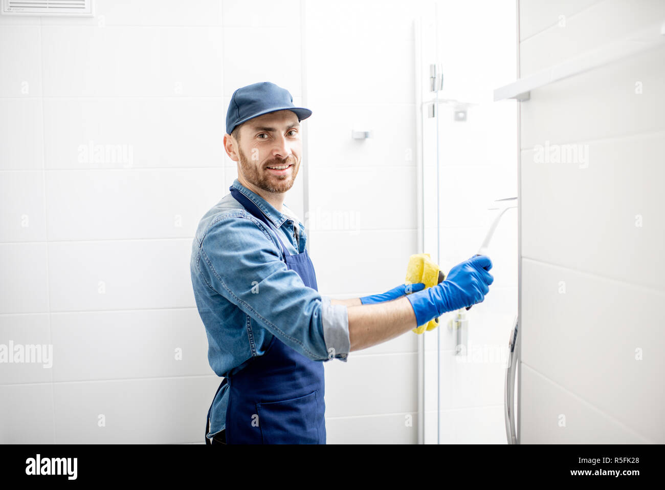 L uomo come pulitore professionale tergi la porta della doccia con tergicristallo di cotone nel bagno bianco Foto Stock