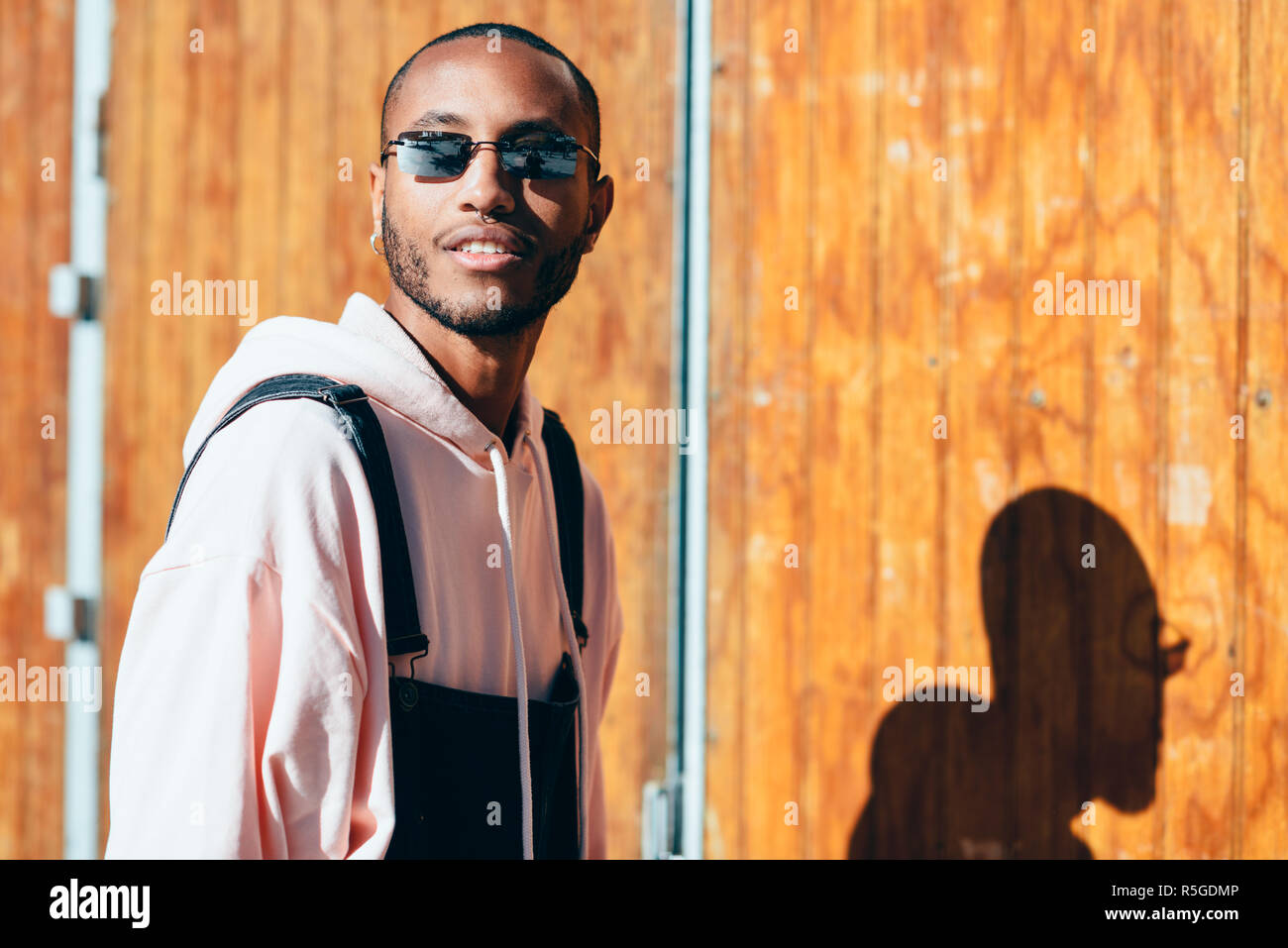 Giovane uomo nero di indossare un abbigliamento informale e occhiali da sole, sorridente contro uno sfondo di legno. Millenaria ragazzo africano con bib pants all'aperto Foto Stock