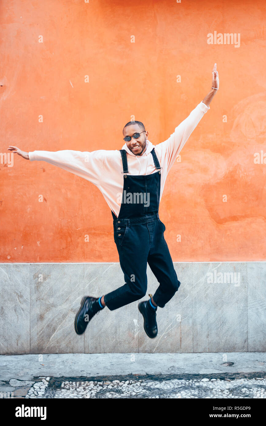Giovane uomo nero indossando abiti casual jumping in background urbano. Il concetto di stile di vita. Millenaria ragazzo africano con bib pants all'aperto Foto Stock