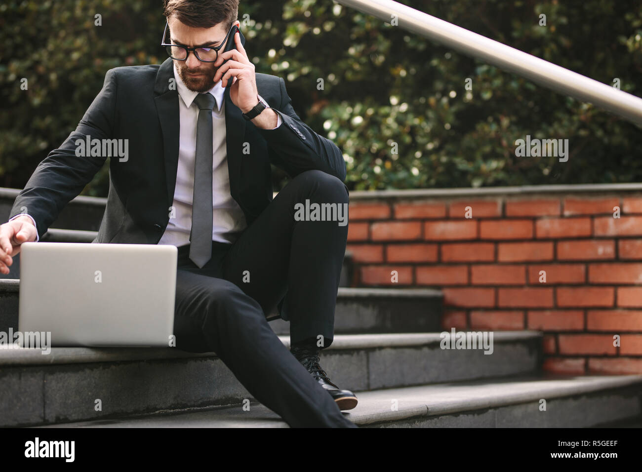 uomo d'affari che lavora su un computer portatile e parla al telefono cellulare mentre è seduto all'aperto sui gradini. Imprenditore maschile che gestisce il lavoro all'aperto. Foto Stock