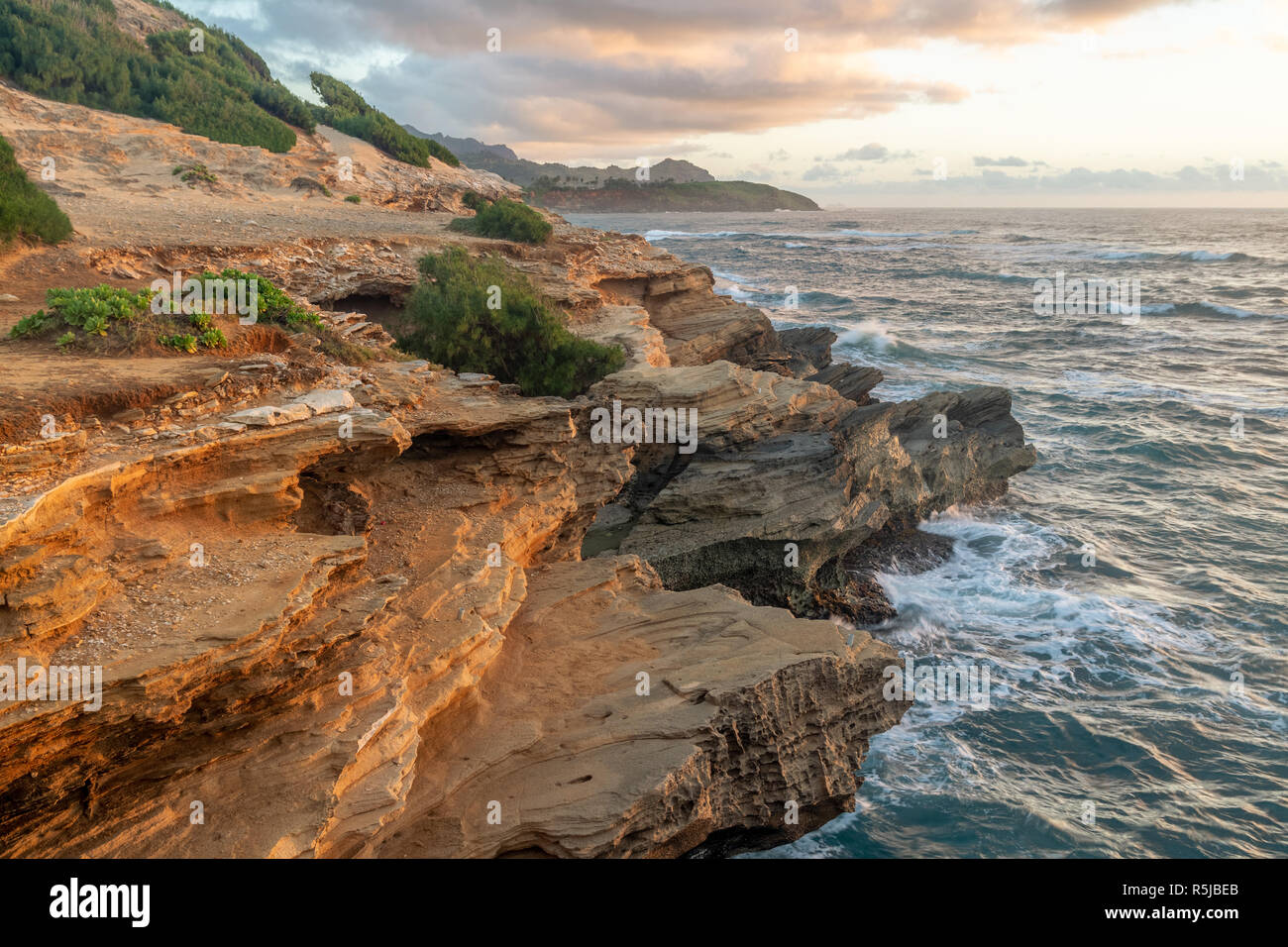 Onde che si infrangono in Makawehi Bluff durante la calda luce alba illumina le nuvole, Kauai, Hawaii Foto Stock