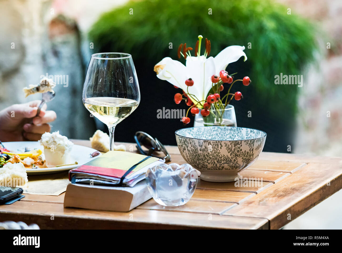 Una donna di mangiare da soli con un bicchiere di vino e un libro e occhiali da sole su di un tavolo di legno, all'esterno. Foto Stock