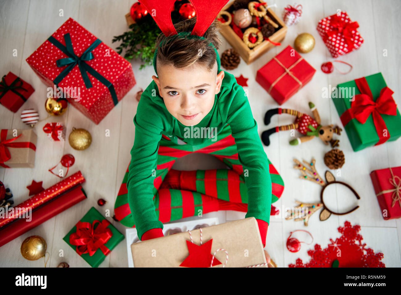 Vista dall'alto di un curioso ragazzo indossa xmas pigiami seduta sul pavimento, tenendo un regalo di Natale, guardando verso l'alto. Foto Stock