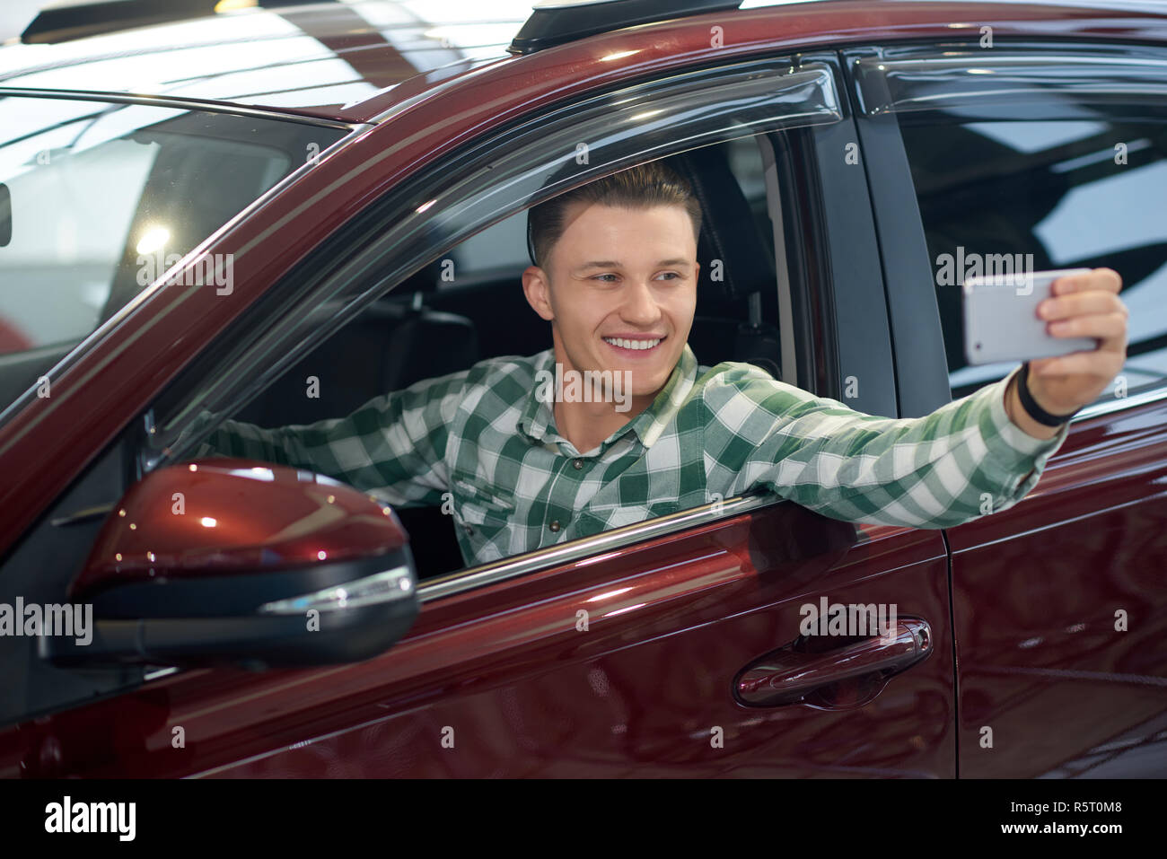 Bella bionda uomo rendendo selfie sul suo telefono e sorridente. Driver attraente in camicia a scacchi seduta in rosso scuro auto. Nuovo felice proprietario di auto di lusso di prendere questo momento sul suo smartphone. Foto Stock