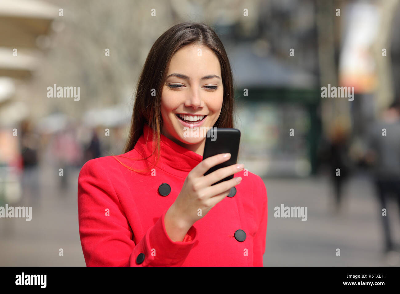 Donna felice indossando giacca rossa passeggiate utilizzando smart phone in inverno in strada Foto Stock