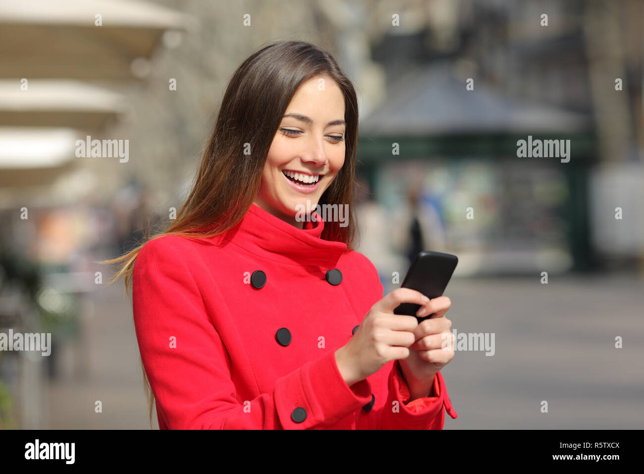 Donna felice indossando giacca rossa in passeggiate invernali e utilizza smart phone in strada Foto Stock