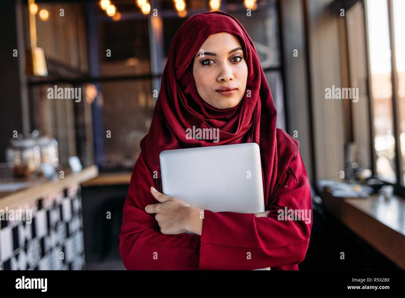 Fiducioso donna in hijab con il computer portatile in piedi al chiuso in un ristorante. Imprenditrice islamica in hijab presso la caffetteria. Foto Stock
